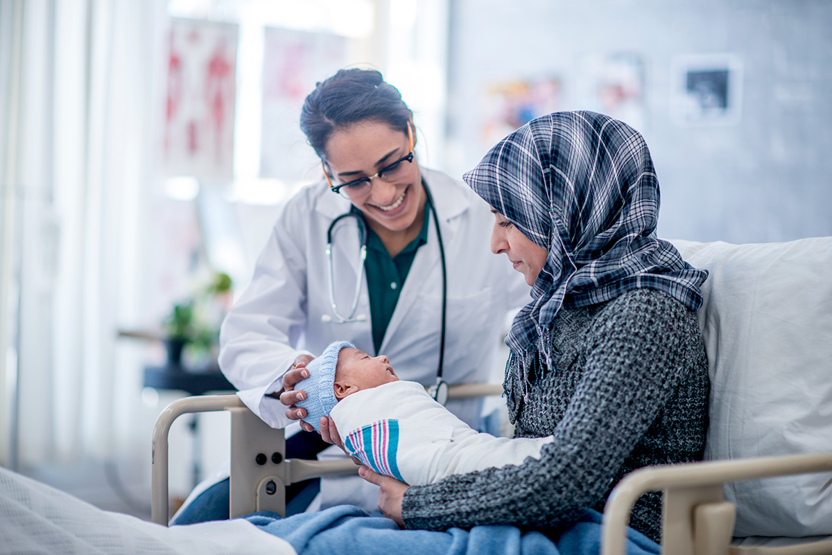 A mother and her newborn baby boy are indoors in a hospital. The mother is holding her baby while laying in the hospital bed. Their doctor is smiling down at the baby.