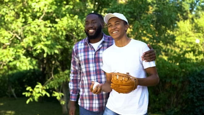 father embracing son wearing white shirt and with baseball glove and ball in hand