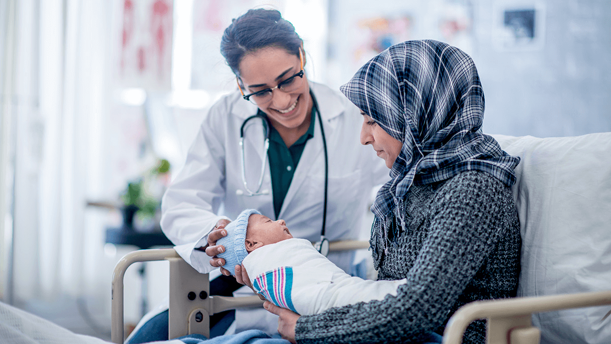 Image of two women seating, one a doctor with white coat and stethoscope and a the other holding a new born baby boy.