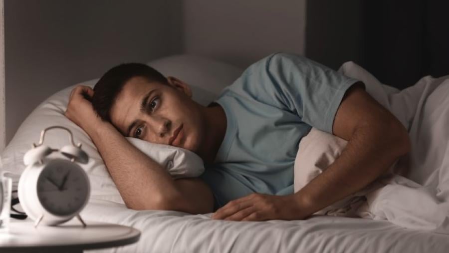 man lying in bed unable to sleep with his clock in the foreground