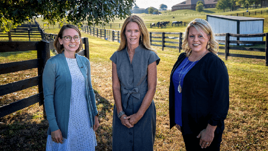 Three women stand next to each other in front of a fence at a horse farm.